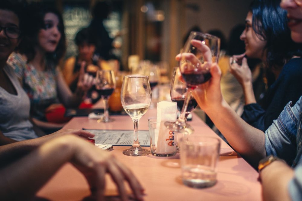Friends leaning over wine glasses at a crowded table, mid-conversation